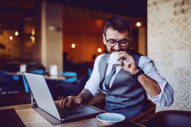 Zadowolony kaukaski przystojny brodaty biznesmen w garniturze siedzi w kawiarni, pije kawę i patrząc na laptopa.