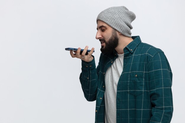 Zadowolony Młody Człowiek W Czapce Zimowej Stojący W Widoku Profilu, Patrzący W Dół, Rozmawiający Przez Telefon Za Pomocą Mikrofonu Na Białym Tle Z Miejscem Na Kopię