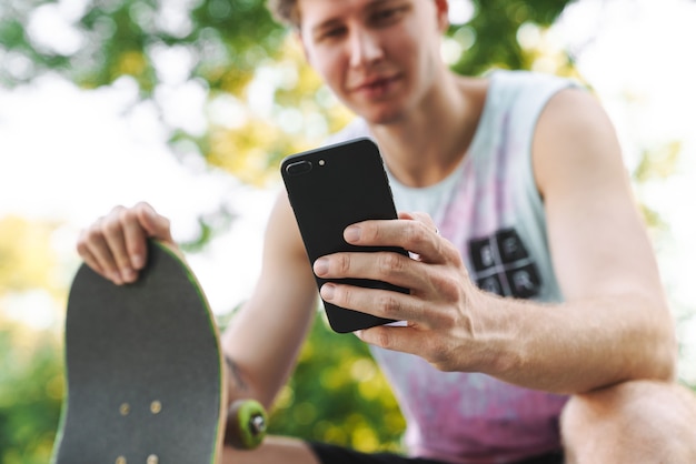 Zdjęcie zadowolony młody człowiek w streetwear siedzący z deskorolką i piszący na telefonie komórkowym w letnim skateparku