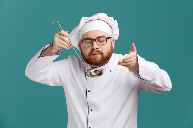Zadowolony młody mężczyzna szef kuchni w mundurze okularowym i czapce trzymającej kadzi trzymającej rękę w powietrzu udaje, że pachnie zapach posiłku z zamkniętymi oczami na białym tle na niebieskim tle