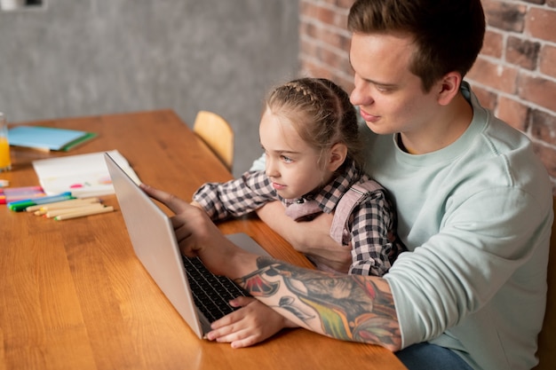 Zadowolony młody ojciec z tatuażem na ramieniu siedzi przy stole i wskazuje na ekran laptopa, wyjaśniając zaciekawionej córce zadanie uczenia się programu
