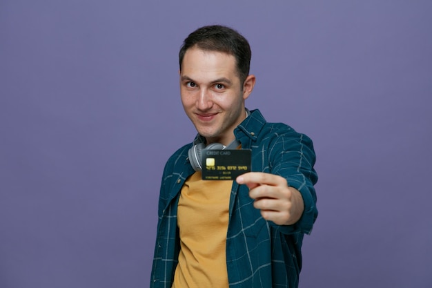 zadowolony młody student noszący słuchawki na szyi stojący w widoku profilu, patrzący na kamerę wyciągającą kartę kredytową w kierunku kamery na fioletowym tle