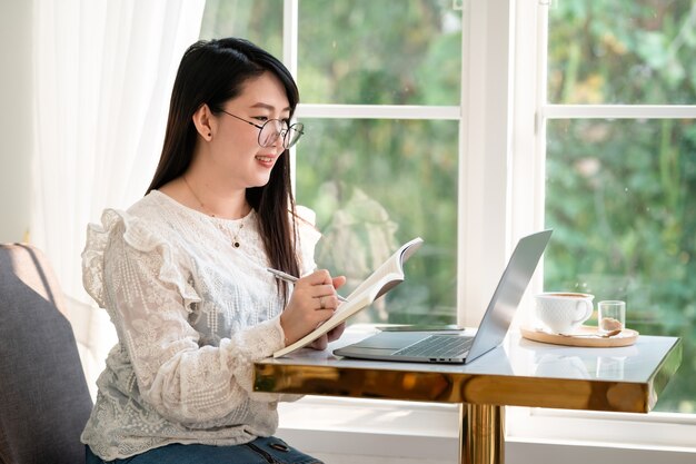 Zadowolony z azjatyckich freelancerów Businesswoman robienia notatek pisemnych na notebooku dorywczo pracując z laptopem z kubkiem na filiżankę kawy i smartfonem w kawiarni, biznesowy styl życia