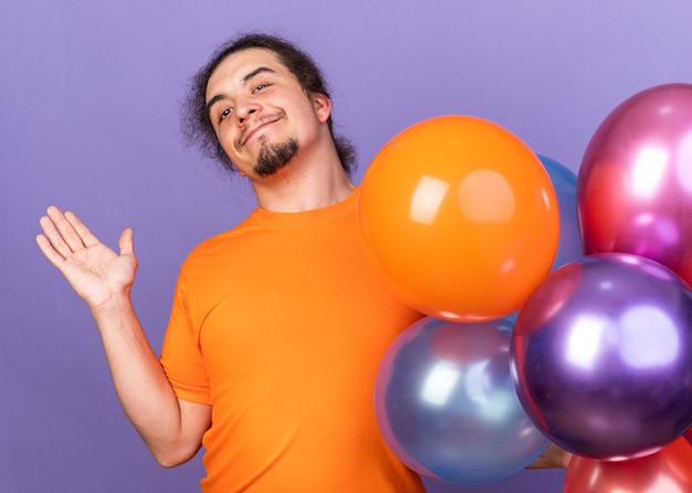 Zadowolony z kamery młody człowiek ubrany w rozłożoną rękę stojącą w pobliżu balonów odizolowanych na fioletowej ścianie