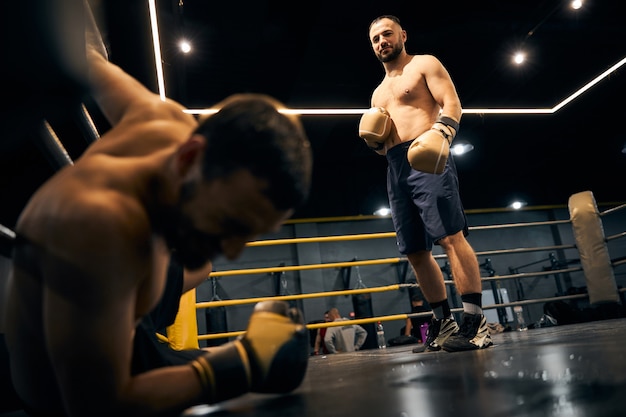 Zadowolony zawodnik stojący na środku ringu patrzący na swojego partnera, leżącego na podłodze w rogu