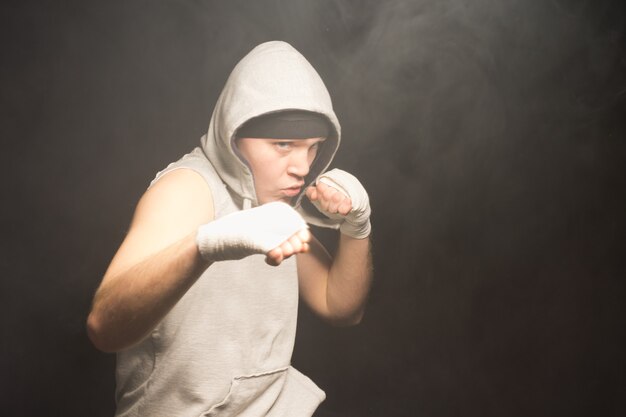 Zadziorny młody bokser w bluzie z kapturem, unoszący zabandażowane pięści w walce w ciemnej, zadymionej atmosferze z copyspace