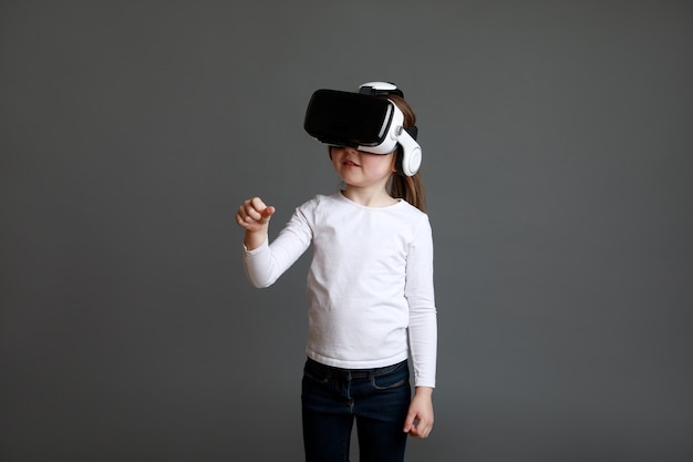 Zafascynowana dziewczynka w białej koszuli z długim rękawem, patrząc w okulary wirtualnej rzeczywistości na szarej powierzchni.