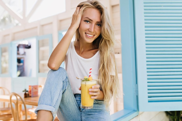 Zainspirowana kobieta z długą fryzurą trzymająca szklankę soku i pozująca z uśmiechem Portret opalonej blond modelki w dżinsach, siedzącej na parapecie i pijącej koktajl