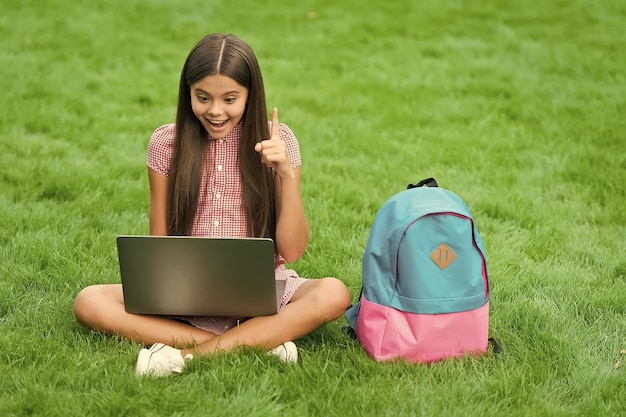 Zainspirowane szczęśliwe dziecko korzystające z komputera do nauki w szkole online, siedzące w parku na zielonej trawie z edukacją plecaka