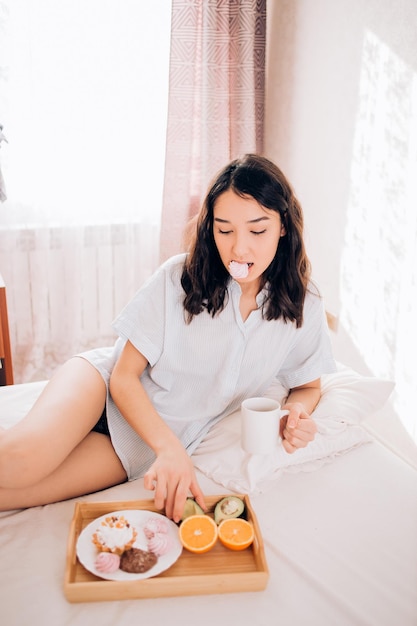 Zainteresowana kaukaska kobieta z gołymi nogami chłodzi rano telefon komórkowy, pijąc kawę. Wewnątrz zdjęcie czarującej dziewczyny siedzącej w łóżku i wysyłającej SMS-a. Śniadanie w łóżku.