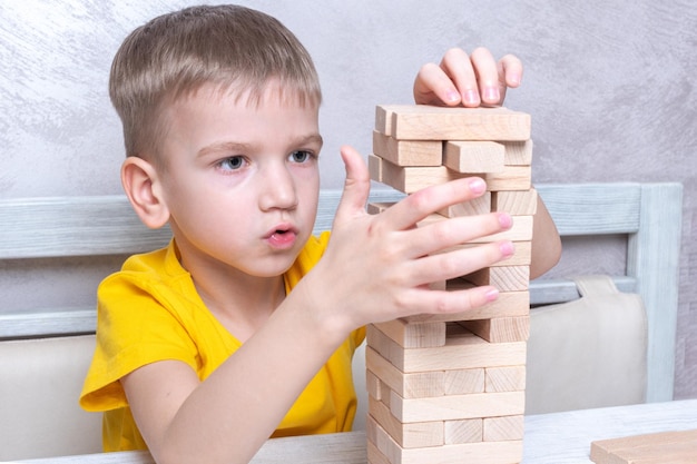 Zdjęcie zainteresowany szczęśliwy mały blond chłopiec grający w grę planszową biorąc klocki z drewnianej wieży utrzymujący równowagę gra planszowa wieża z drewnianych klocków aktywność dla strategii zwinności koncentracji