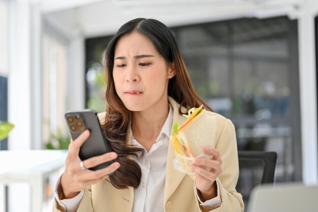 Zajęta i głodna azjatycka bizneswoman je kanapkę, odpowiadając na e-maile przez telefon