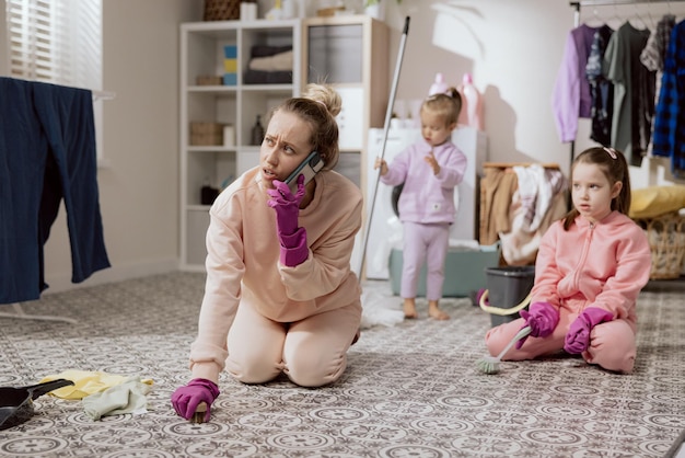 Zajęta kobieta ma gumowe różowe rękawiczki na rękach Matka dwóch córek czyści łazienkę na kolanach szorując podłogę szczotką do polerowania i rozmawia przez telefon Małe dziewczynki pomagają mamie sprzątać