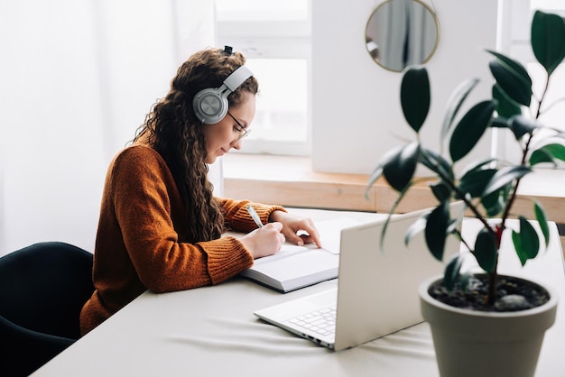 Zajęta młoda kobieta, studentka uniwersytetu, nosząca słuchawki za pomocą laptopa e-learning, pisząca notatki, studiując