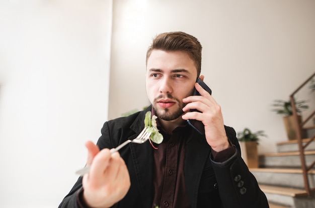 Zajęty biznesmen siedzi w przytulnej restauracji, trzymając widelec z jedzeniem i rozmawia przez telefon