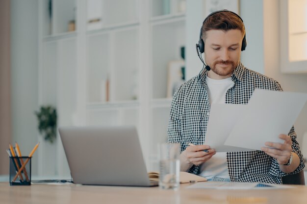 Zajęty mężczyzna freelancer noszący zestaw słuchawkowy, trzymający dokumenty w rękach i pracujący zdalnie na laptopie, siedząc w salonie w domu. Koncepcja biznesowa online i freelancer