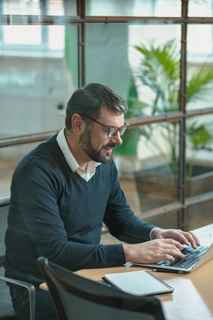 Zajęty mężczyzna siedzi przy biurku i pracuje na laptopie podczas pisania