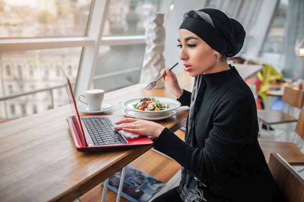 Zajęty młoda kobieta arabska pracy na laptopie i spójrz na to. Siedzi w środku i trzyma widelec na miskach z jedzeniem. Model nosi hidżab.