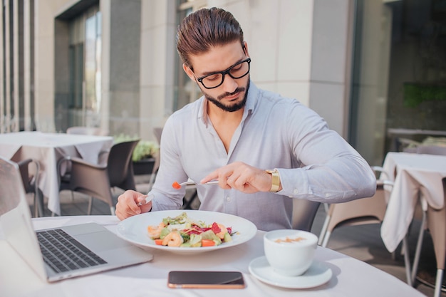 Zajęty młody człowiek siedzi przy stole na zewnątrz restauracji. Patrzy na zegarki. Przy stole jest laptop, miska sałatki, telefon i filiżanka kawy.