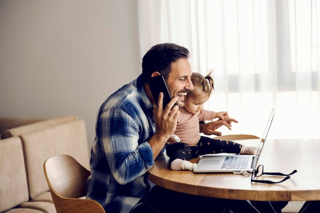 Zajęty ojciec, który zostaje w domu, rozmawia przez telefon z pracy, uśmiechając się do laptopa