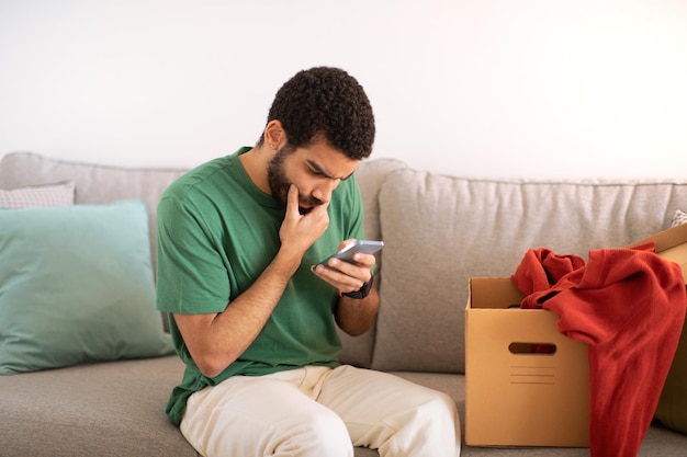 Zajęty, zamyślony, młody bloger z Bliskiego Wschodu z kartonowym pudełkiem patrzy na smartfona