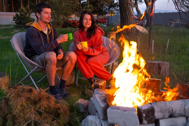 Zakochana para siedzi przy ognisku na podwórku domu na krzesłach. Romantyczny wieczór. Mężczyzna i kobieta ogrzewają ręce przy ogniu, piją herbatę.