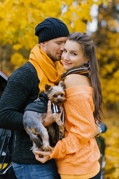 Zakochany facet i dziewczyna przytulają się w jesiennym parku, facet trzyma w ramionach uroczego psa. Portret młodej rodziny ze zwierzakiem