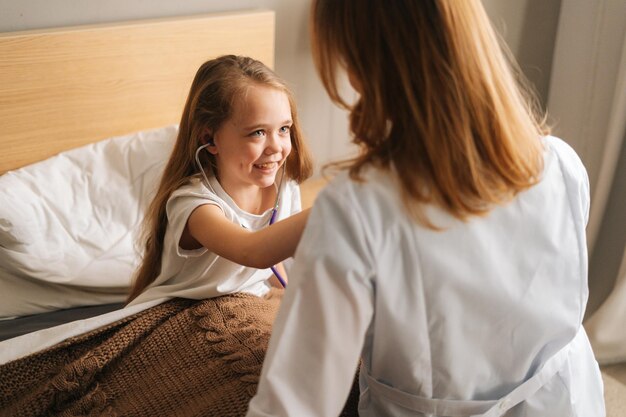 Zakończenie szczęśliwa mała dziewczynka słucha bicie serca stetoskopem do lekarki siedzącej w łóżku. Słodkie dziecko grając jako lekarz pielęgniarka słuchający lekarz ze stetoskopem. Pojęcie zdrowia dzieci