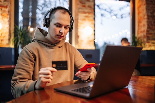 Zakupy I Płatność Online Za Pomocą Telefonu Komórkowego I Karty Kredytowej. Mężczyzna Siedzący W Kawiarni, Ubrany W Zwykły Strój, Kupuje Towary I Płaci Przez Internet, Używając Czarnej Karty Debetowej. Nowa Koncepcja Normalności I Technologii.