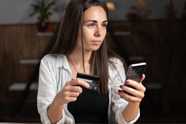 Zakupy online Dziewczyna płaci telefonem komórkowym i kartą kredytową siedząc przy stole w domu Bezgotówkowa płatność za zamówienie w Internecie Młoda kobieta korzysta z technologii