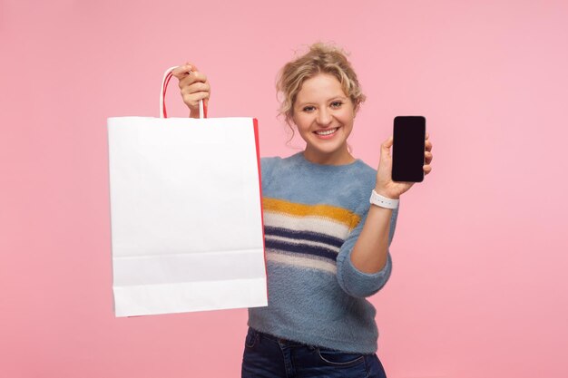 Zakupy online. Portret szczęśliwa dorosła kobieta z kręconymi włosami w swetrze, trzymając telefon komórkowy i torby na zakupy, patrząc na kamerę z uroczym uśmiechem. strzał w studio na białym tle na różowym tle