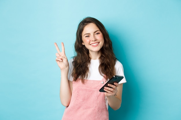 Zakupy online. Śliczna i wesoła młoda kobieta uśmiecha się, trzyma smartfon i pokazuje znak v pokoju przed kamerą, stojąc na niebieskim tle.