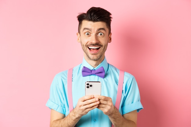 Zakupy online. Szczęśliwy człowiek patrząc zdziwiony po przeczytaniu ekranu smartfona, uśmiechając się podekscytowany do kamery, stojąc na różowym tle.