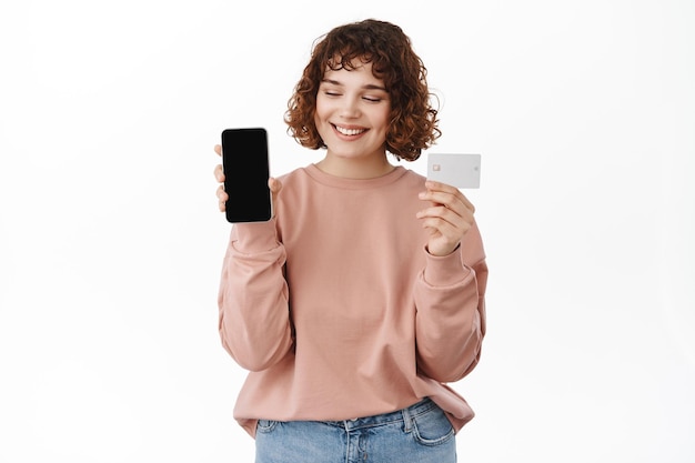 Zakupy online. Uśmiechnięta kędzierzawa dziewczyna patrzy na ekran swojego telefonu komórkowego, pokazując ekran smartfona i kartę kredytową z zadowoloną twarzą, stojąc na białym tle
