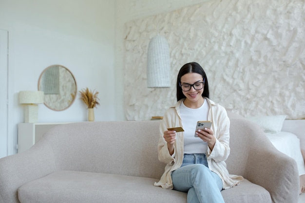 Zakupy online zadowolona młoda piękna kobieta w okularach siedząca na kanapie w domu posiada kredyt