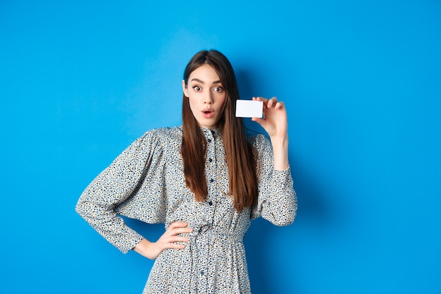 Zakupy. Podekscytowana dziewczyna pokazuje swoją plastikową kartę kredytową i patrzy na zniżki, mówi wow zdziwiona, stojąc na niebiesko.