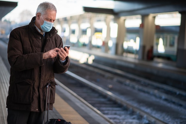 Zamaskowany biznesmen korzystający ze smartfona podczas oczekiwania na koncepcję mobilności w pociągu i koronawirusie