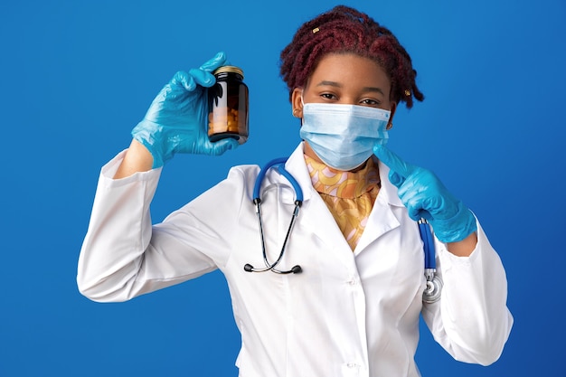 Zamaskowany lekarz afroamerykanin pokazujący szklany słoik leków na niebieskim tle