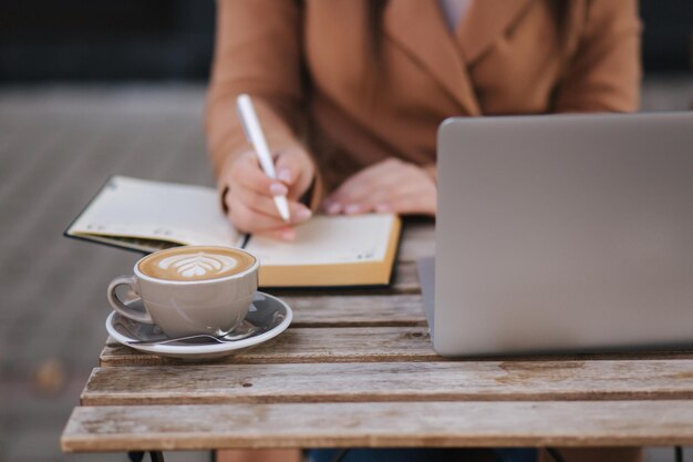 Zamknąć procesu roboczego w kawiarni na zewnątrz Kobieta spisując coś w notatniku Cappuccino laptop i kalendarz na stole