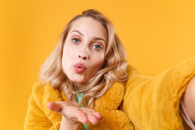 Zdjęcie zamknij się całkiem młoda blond kobieta w żółtym futrze pozowanie na białym tle na pomarańczowym tle. koncepcja życia ludzi. makieta miejsca na kopię. robi selfie strzał na telefon komórkowy dmuchanie wysyłając pocałunek powietrza.