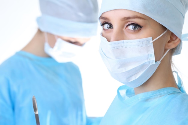Zamknij Się Kobieta Chirurg Patrząc Na Kamery, Podczas Gdy Koledzy Występujący W Tle W Sali Operacyjnej, Na Białym Tle. Koncepcja Chirurgii I Medycyny