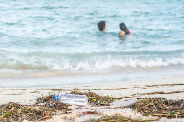 Zamknij się obraz pustej plastikowej butelki z wodą na brudnej plaży wypełnionej wodorostami, śmieciami i odpadami na brudnej, piaszczystej plaży z ludźmi na morzu