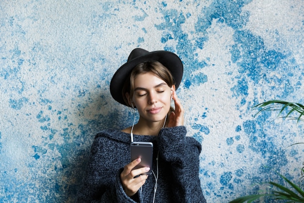 Zamknij Się Portret Młodej Kobiety Ubranej W Sweter I Kapelusz Na Niebieskiej ścianie, Słuchając Muzyki Przez Słuchawki, Trzymając Telefon Komórkowy, Oczy Zamknięte