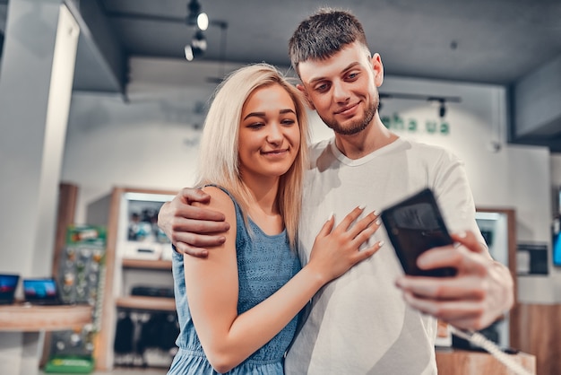 Zamknij się widok ostrości stylowej, uroczej, obejmującej młodych studentów miłości, biorąc selfie z nowym telefonem komórkowym w sklepie technologicznym.