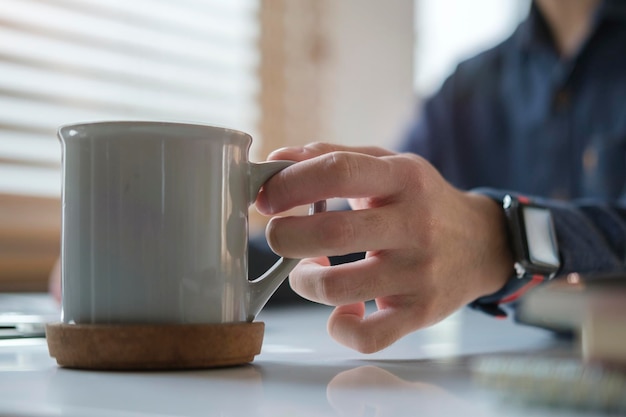 Zamknij widok człowieka ręki trzymającej filiżankę kawy nad białym stołem w pobliżu okna w nowoczesnym biurze domowym