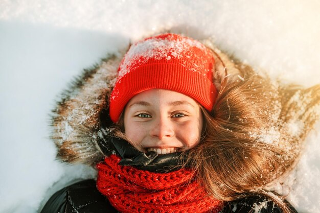 Zamknij zimowy portret uśmiechniętej dziewczynki w czerwonym kapeluszu leżącej w śnieżnym widoku z góry