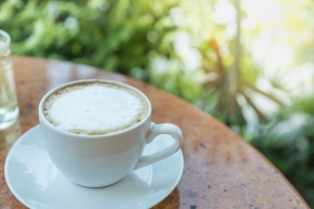 Zamyka up biała filiżanka i talerz gorąca latte kawa na round stole i zieleni liścia naturze