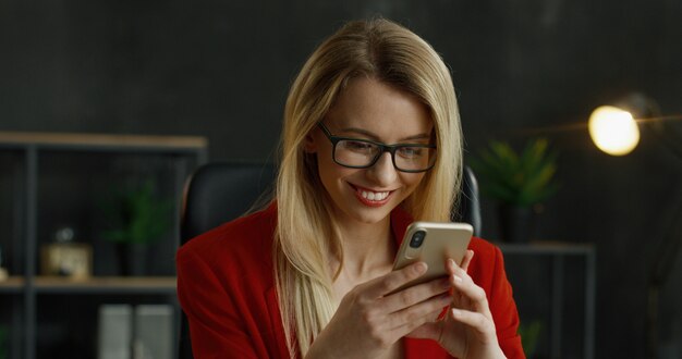 Zamyka up smartphone w rękach blondynka bizneswoman. Szczęśliwa żeńska wiadomość tekstowa i pisać na telefonie komórkowym w gabinecie.