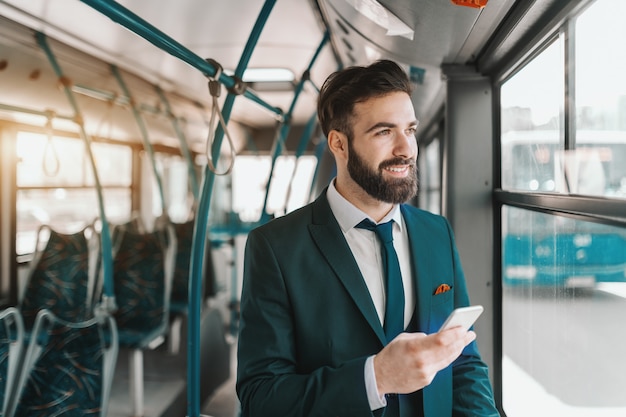 Zamyka up uśmiechnięty brodaty biznesmen w formalnej odzieży używać mądrze telefon i patrzejący przez okno podczas gdy stojący publicznie transport.
