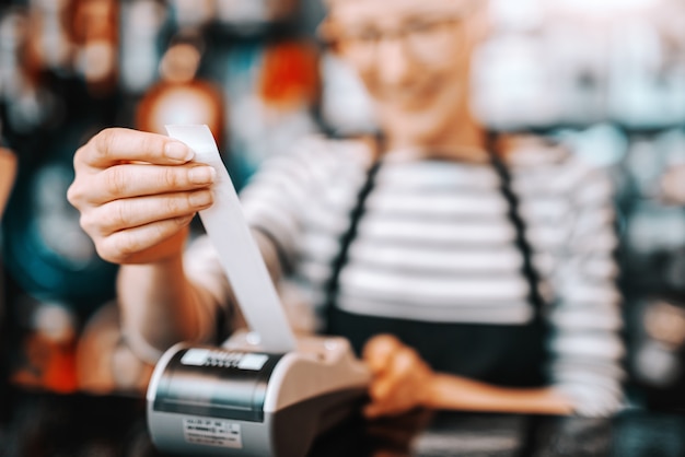 Zdjęcie zamyka up uśmiechnięty kaukaski żeński pracownik z krótkim blondynka włosy i eyeglasses używać kasę podczas gdy stojący w rowerowym sklepie.